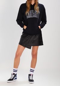 Svart huvtröja med "Carhartt"-logga, kombinerad med en svart konstläder-wrapkjol, vita strumpor med marinblå ränder och hög topp sneakers.