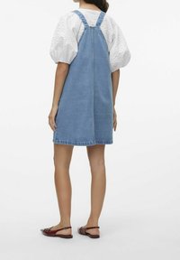 Kvinna som bär en blå denimpinafore klänning över en vit blus med puffärm och bruna platta slingback-skor, stående mot en enfärgad bakgrund.