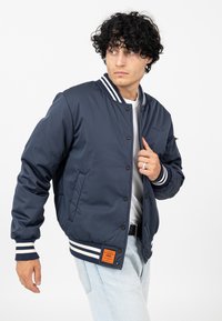 Marineblauwe bomberjack van gladde stof heeft witte gestreepte ribgebreide manchetten en kraag, met een zijzak en een oranje labeldetail.