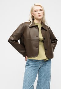 Young woman with long blonde hair wearing a brown leather jacket, olive green top, and blue patterned wide-leg pants against a plain background.