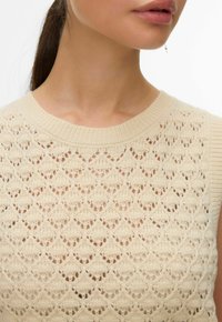 Femme portant un haut beige en maille sans manches avec un motif ajouré complexe, montrant le cou, le menton et les lèvres en gros plan.