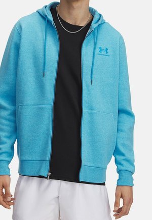 Mężczyzna w jasnoniebieskiej bluzie z kapturem na zamek marki Under Armour, na czarnej koszulce i białych szortach, pokazany od ramion do bioder.