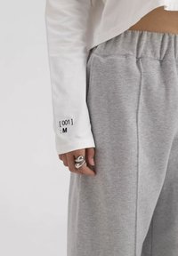 Witte lange mouw top met geprinte details op de manchetten, gecombineerd met grijze sweatpants. Zilveren ring gedragen aan een hand die op de broek rust.
