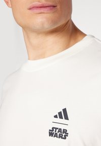 Λευκό βαμβακερό T-shirt με στρογγυλή λαιμόκοψη, με μαύρο λογότυπο Adidas και γραφικό "STAR WARS" τυπωμένο στο στήθος. Λείος υφασμάτινος χαρακτήρας.