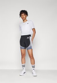 Hvit crop top med svart logo, svarte og grå tettsittende shortser med buede mønstre, kombinert med stripete sokker og hvite joggesko.