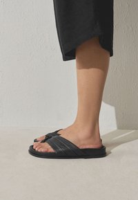 Personne portant des sandales noires à larges brides croisées et un pantalon noir raccourci, debout sur un sol clair.