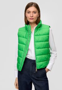 Grüne gepolsterte Weste mit hohem Kragen und Reißverschluss. Hat eine gesteppten Struktur und glänzende Oberfläche. Trägt man über einem weißen Hemd und dunklen Hosen.