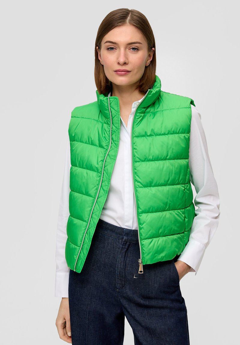 Grüne gepolsterte Weste mit hohem Kragen und Reißverschluss. Hat eine gesteppten Struktur und glänzende Oberfläche. Trägt man über einem weißen Hemd und dunklen Hosen.