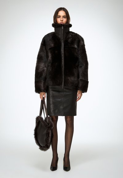 Veste en fausse fourrure noire à col montant, fermeture éclair et grandes poches, associée à une jupe en cuir noir ajustée et un sac assorti.
