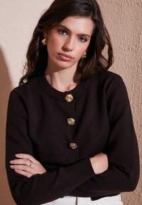 Tmavo hnedý pletený cardigan s okrúhlym výstrihom, ktorý má štyri zlaté gombíky v tvare kvetu a rebrované manžety. Hladká textúra, priliehavý strih.