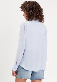 Chemise à rayures bleu clair à manches longues avec une coupe décontractée, dotée d'un ourlet arrondi et de fentes au niveau des poignets avec boutons, associée à un short en denim délavé.