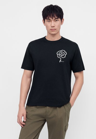 Jeune homme aux cheveux courts et noirs porte un t-shirt noir avec un motif spirale abstrait blanc sur la poitrine gauche et un pantalon vert, main dans la poche.