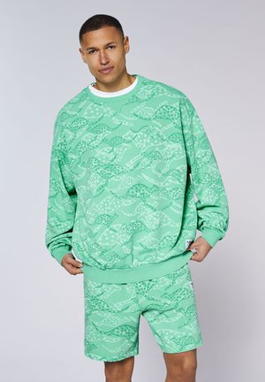 Man wearing matching mint green sweatshirt and shorts with abstract wavy dot patterns, standing against plain background.
