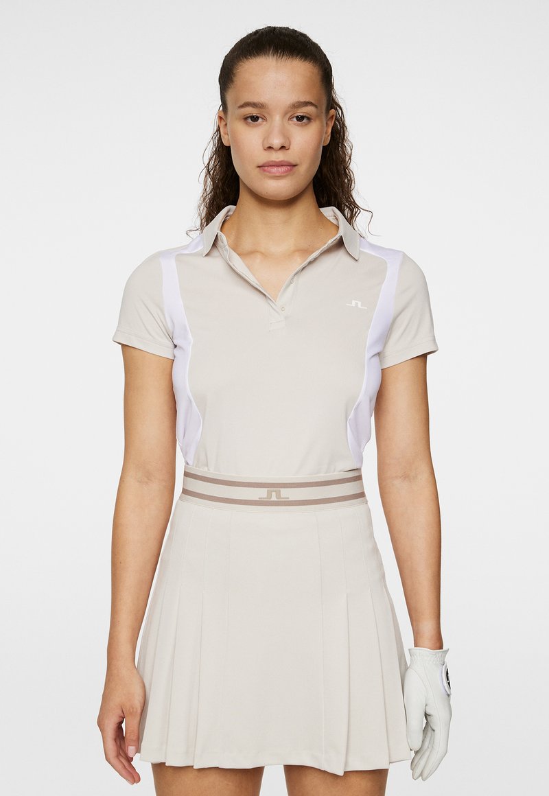 Beige golfpolo med vita sidodetaljer, kort ärm och krage. Matchas med en beige plisserad kjol och en vit golfhandske.
