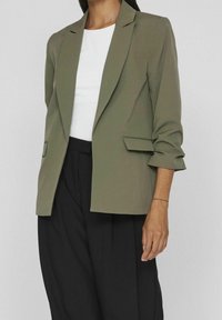 Blazer vert olive avec une coupe ajustée, des revers crantés, deux poches avant et des manches retroussées, porté avec un haut blanc et un pantalon noir.
