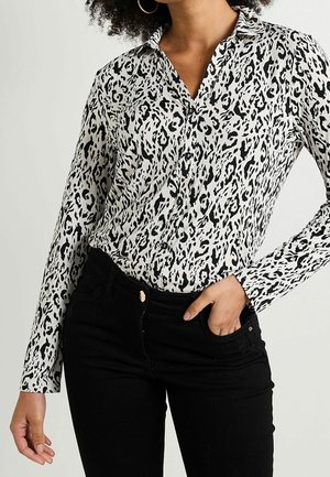 Vrouw draagt een zwart-wit leopardprint blouse met lange mouwen en knopen en een zwarte broek, één hand in de zak, tegen een effen achtergrond.