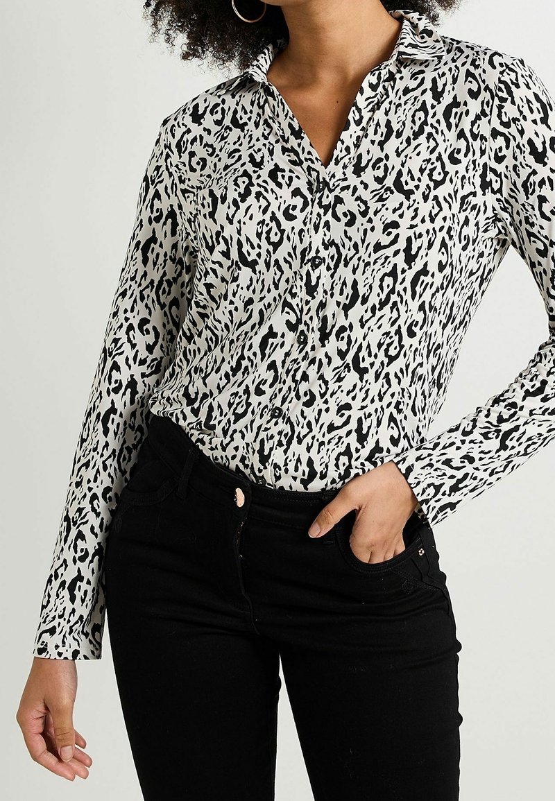 Femme portant une chemise à manches longues à boutons avec imprimé léopard noir et blanc et un pantalon noir, une main dans la poche, sur un fond uni.