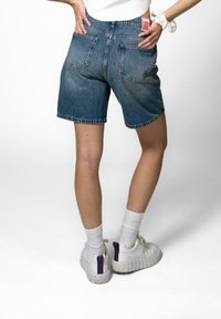 Denimshorts i medium blå med regular fit, med høy midje, oppbrettede hem og to baklommer. Brukt med hvite sneakers og sokker.