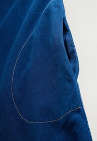 Țesătură din denim albastru cu o textură netedă, având o cusătură curbată conturată cu fir deschis. Țesătura prezintă o ușoară strălucire.
