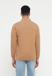 Barna bordázott pulóver, gallérral és hosszú ujjakkal. Az anyag texturált felületű. Világoskék farmernadrággal párosítva.