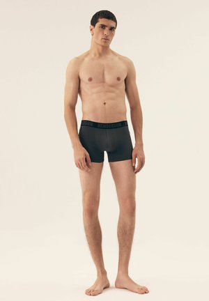 Schwarze Boxershorts mit einem elastischen Bündchen, das mit einer Marke versehen ist, getragen von einem oberkörperfreien männlichen Modell, das ein passgenaues Design und eine glatte Textur präsentiert.