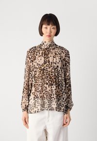 Femme aux cheveux courts et foncés portant un blouse à imprimé léopard à col haut et manches longues, ainsi que des pantalons blancs, sur un fond uni.