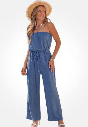 Mujer con un mono azul sin tirantes y sombrero de paja, complementada con collares en capas, pulseras, anillos y sandalias blancas de tacón alto.