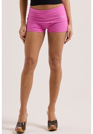 Frau trägt pinke Shorts und schwarze Plateau-Sandalen, steht vor einem schlichten hellen Hintergrund.