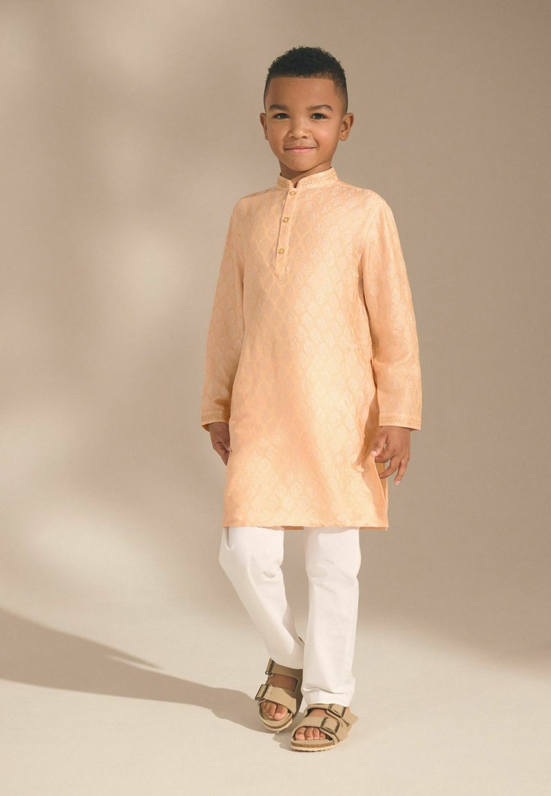 Junger Junge trägt eine pfirsichfarbene Langarm-Kurta mit weißen Hosen und braunen Sandalen, steht vor einem schlichten beigen Hintergrund.