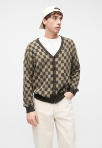 Νεαρός άνδρας που φοράει ένα μπεζ και σκούρο πράσινο καρό ζακέτα πάνω από λευκό πουκάμισο, κρεμ παντελόνι και μπεζ καπέλο φοράει ανάποδα, κοιτάζοντας προς τα δεξιά.