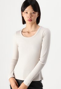 Long-sleeve cream ribbed top with a scoop neckline, featuring a fitted silhouette and smooth texture. Styled with black pants.