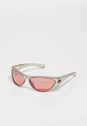Lunettes de soleil avec des verres teintés roses et une monture blanche, ornées d'accents rouges et de détails découpés sur les côtés, offrant une apparence sportive.
