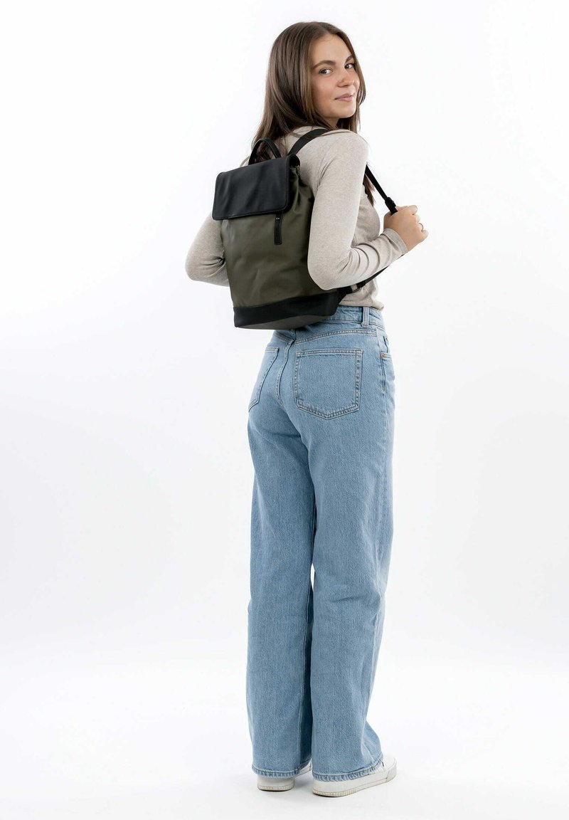 Rucksack aus grünem Stoff mit schwarzem Oberteil, ausgestattet mit verstellbaren Trägern und einer seitlichen Reißverschlusstasche. Modell trägt hellblaue Jeans.
