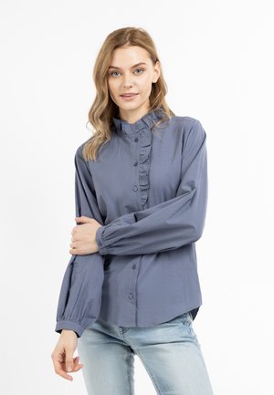 Frau mit hellbraunen Haaren, die eine langärmelige blaue Bluse mit Knöpfen und Rüschen sowie hellblaue Jeans trägt, steht vor einem weißen Hintergrund.