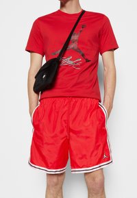 Czerwona bawełniana koszulka z czarnym i srebrnym nadrukiem graficznym; zestawiona z błyszczącymi czerwonymi szortami z białymi paskami po bokach i akcentem logo.