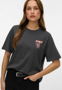 Ciemnoszara bawełniana koszulka z krótkimi rękawami. Na lewej piersi znajduje się różowa grafika w stylu kreskówkowym oraz tekst. Połączona z czarnymi spodniami i paskiem.