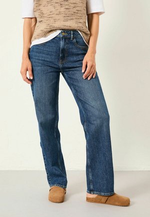 Person trägt blaue Jeans mit geradem Bein, ein weißes Hemd, eine beige Strickweste und beige Slipper und steht vor einem schlichten Hintergrund.