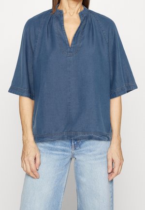 Person, die ein locker sitzendes dunkelblaues kurzärmeliges Oberteil mit V-Ausschnitt und hellblaue Jeans trägt, vor einem schlichten Hintergrund steht.