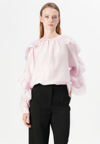 Blusa rosa chiaro con maniche a volant, scollatura rotonda e finitura testurizzata; abbinata a pantaloni neri a vita alta.