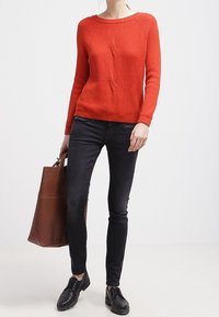 Suéter rojo acanalado con un diseño ligeramente retorcido, combinado con jeans ajustados negros y zapatos negros con cordones; lleva un bolso tote marrón.
