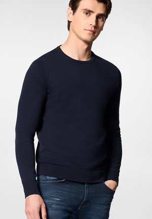Granatowy sweter dzianinowy z okrągłym dekoltem, długimi rękawami oraz ściągaczami u mankietów i na dolnym brzegu. Noszony z przetartymi niebieskimi dżinsami.