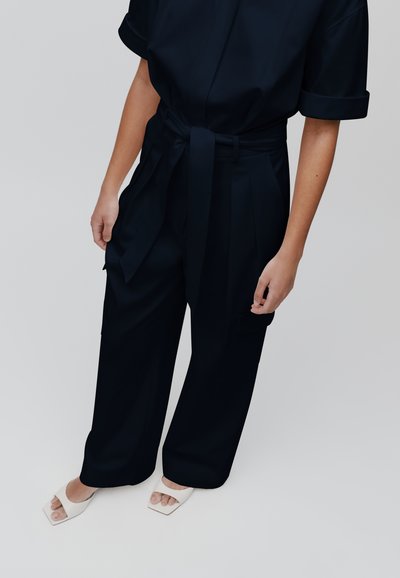 Mono azul marino con mangas cortas y cintura atada, con piernas anchas y bolsillos laterales, combinado con sandalias blancas de punta cuadrada.