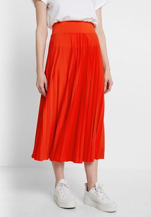 Plisséhouten oranje midi-rok met een hoge tailleband; gedragen met een wit t-shirt en witte sneakers, met een gladde stof en een vloeiende design.