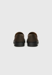 Zapatos marrones de tipo slip-on con un material suave, punta redonda y una suela de goma negra gruesa. El diseño presenta detalles de costura sutiles.