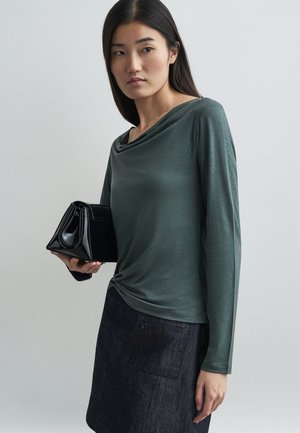 Halenka s dlhými rukávmi a golierom v ztlmené zelenej farbe. Spárovaná s čiernou, štruktúrovanou kabelkou. Pozadie je neutrálne a jemné.