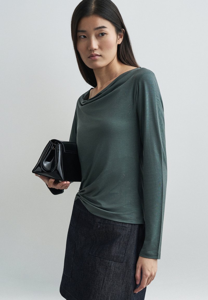 Blusa de manga longa com gola drapeada em tecido verde suave. Acompanhada de uma bolsa estruturada preta. O fundo é neutro e suave.