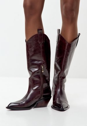 Bottes de cowboy en cuir marron avec un bout pointu, hauteur jusqu'à la cheville, coutures décoratives, fermeture éclair latérale et un talon compensé empilé.