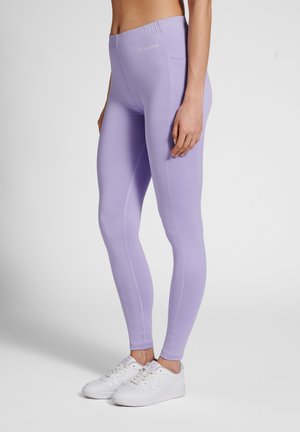 Kvinde iført lyse lilla leggings med høj talje og sidelomme samt hvide sneakers, stående mod en ensartet baggrund.