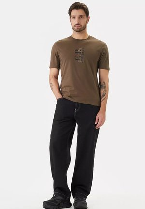 Uomo con tatuaggi che indossa una t-shirt grafica marrone, pantaloni neri a gamba larga e scarpe nere, in piedi con una mano in tasca.
