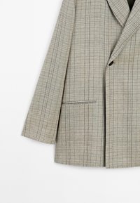 Blazer gris clair à carreaux avec un bouton noir unique, col à revers cranté et poche frontale horizontale sur fond blanc.